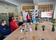 Mehrere Personen sitzen und stehen in einem Konferenzraum mit Bücherregalen. Eine Frau präsentiert vor einem Bildschirm und einem Flipchart mit der Aufschrift „Herzlich willkommen“. Auf dem Tisch stehen Pflanzen, Snacks und Schreibmaterialien.