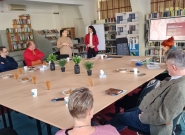 Menschen sitzen in einem Konferenzraum mit Bücherregalen. Zwei Frauen stehen vorne und sprechen, ein Bildschirm zeigt eine Präsentation. Auf dem Tisch stehen Pflanzen, Snacks und Schreibmaterialien. Die Atmosphäre ist konzentriert und professionell.