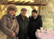 Drei Personen in Winterkleidung stehen lächelnd an einem Stehtisch mit Punschbechern. Im Hintergrund sind eine hölzerne Überdachung und eine Steinmauer zu sehen, die an einen weihnachtlich dekorierten Adventmarkt erinnern.