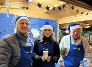 Drei Personen in blauen Rotary-Schürzen und Weihnachtsmützen stehen an einem festlich geschmückten Stand mit goldenen Sternen und blauer Dekoration. Sie halten Punschbecher und lächeln in die Kamera.